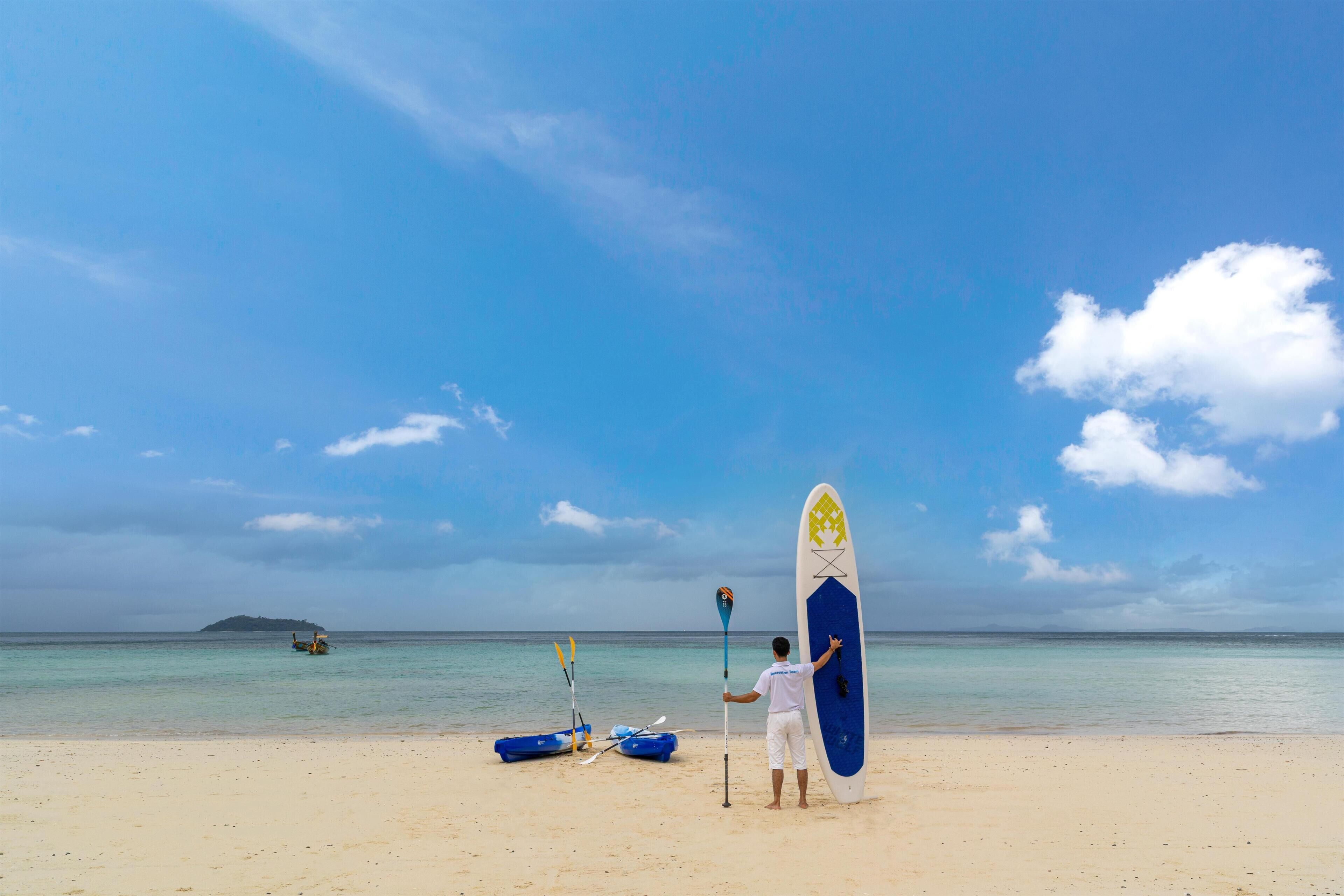A beach scene with a man holding a paddleboard, kayaks, and a paddle on the sand.