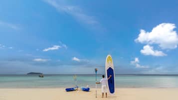 A beach scene with a man holding a paddleboard, kayaks, and a paddle on the sand.