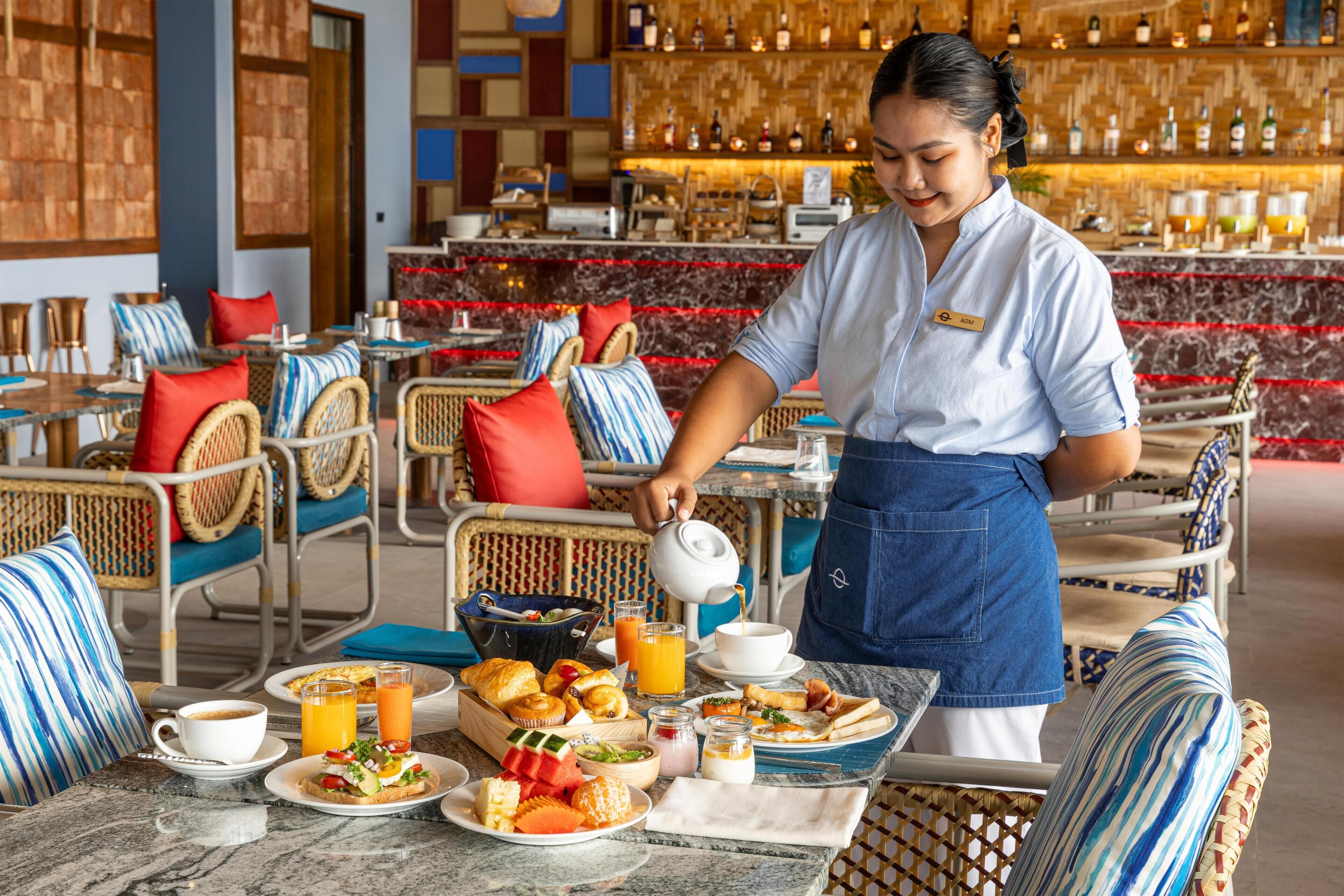 A hotel dining area with a bar in the background, wicker furniture, and a person serving coffee.