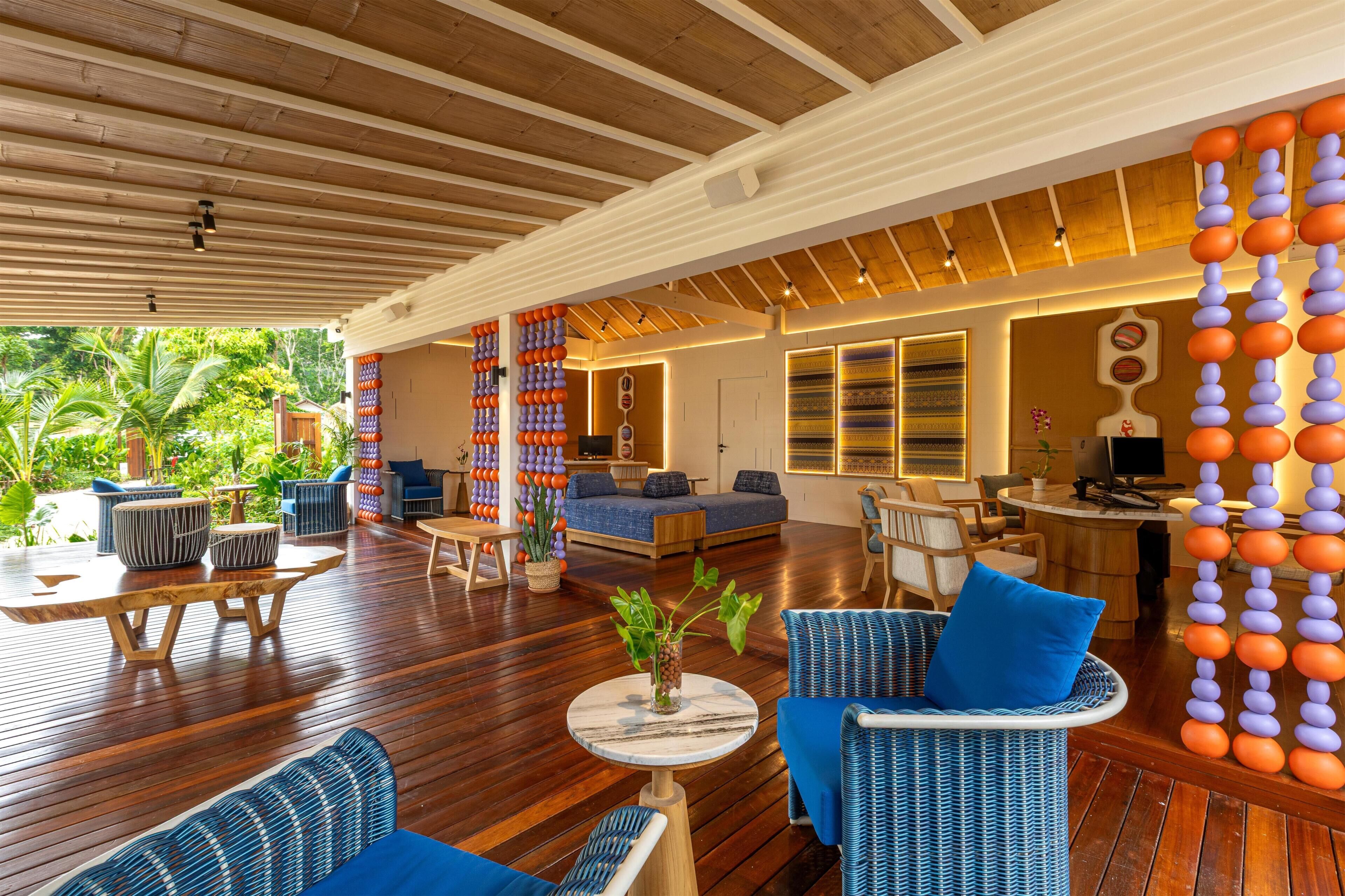 A spacious indoor area with wooden flooring, blue and white wicker furniture, and a large window overlooking a tropical garden.