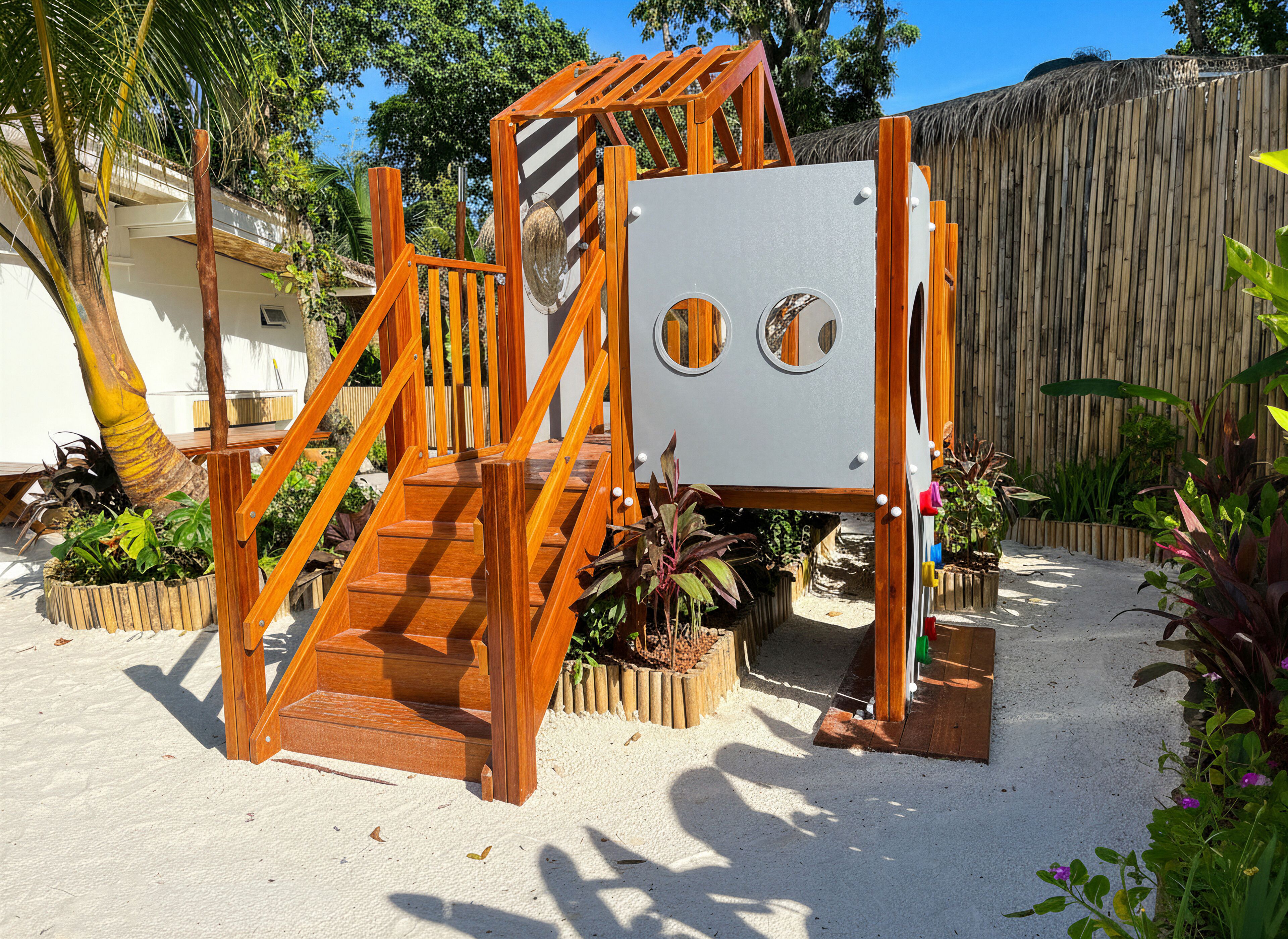 A wooden play structure with a slide and a climbing wall, surrounded by plants and a bamboo fence.