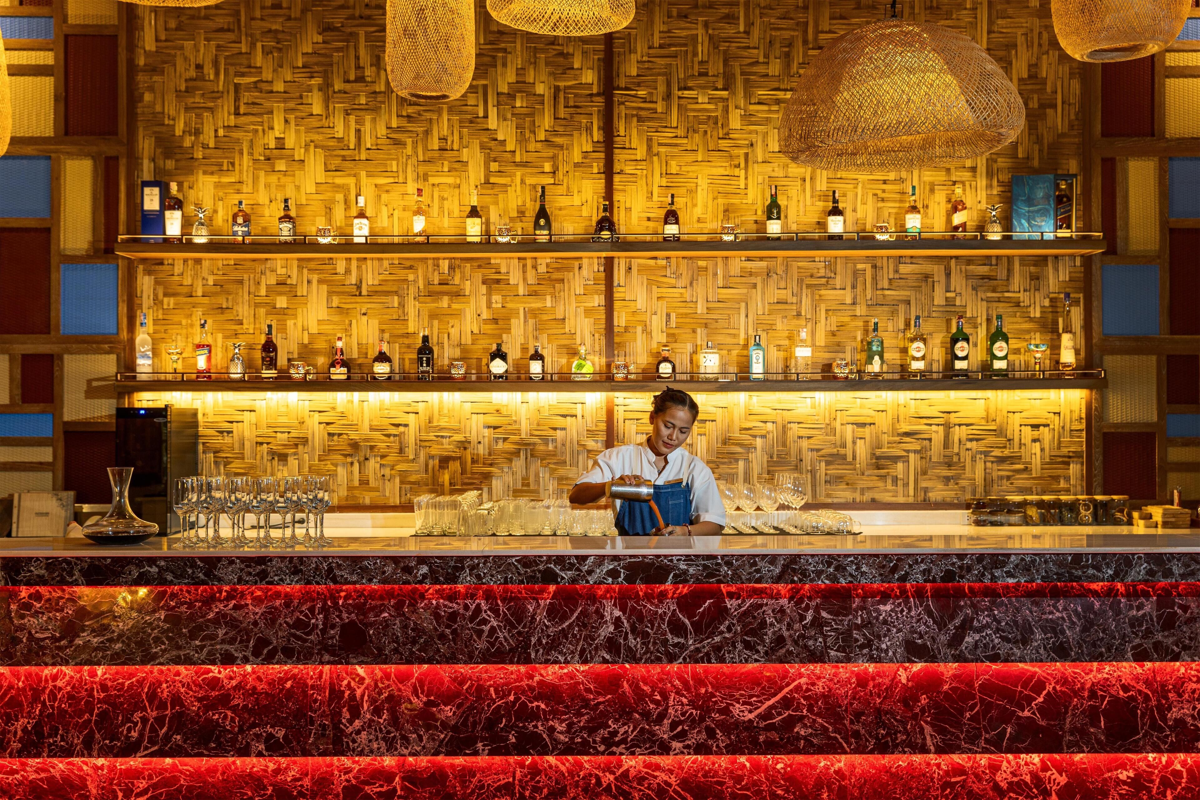 A bar area with a bartender preparing drinks, shelves of liquor bottles, and a woven wall design.