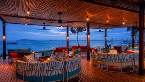 Un espacio de descanso al aire libre con mobiliario de mimbre y cojines rojos, con vistas a un mar tranquilo con barcos.