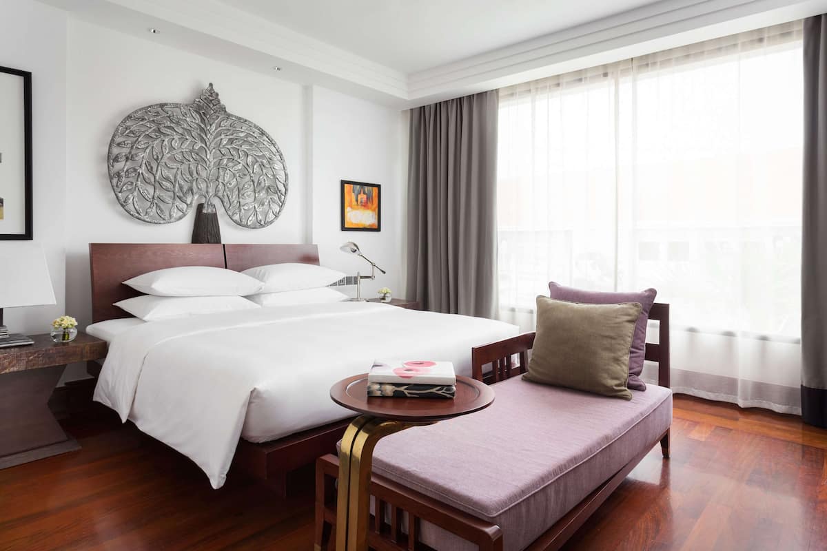Hotelkamer met een groot bed, een bank, een klein tafeltje met een boek, een decoratief wandobject en een groot raam met gordijnen.