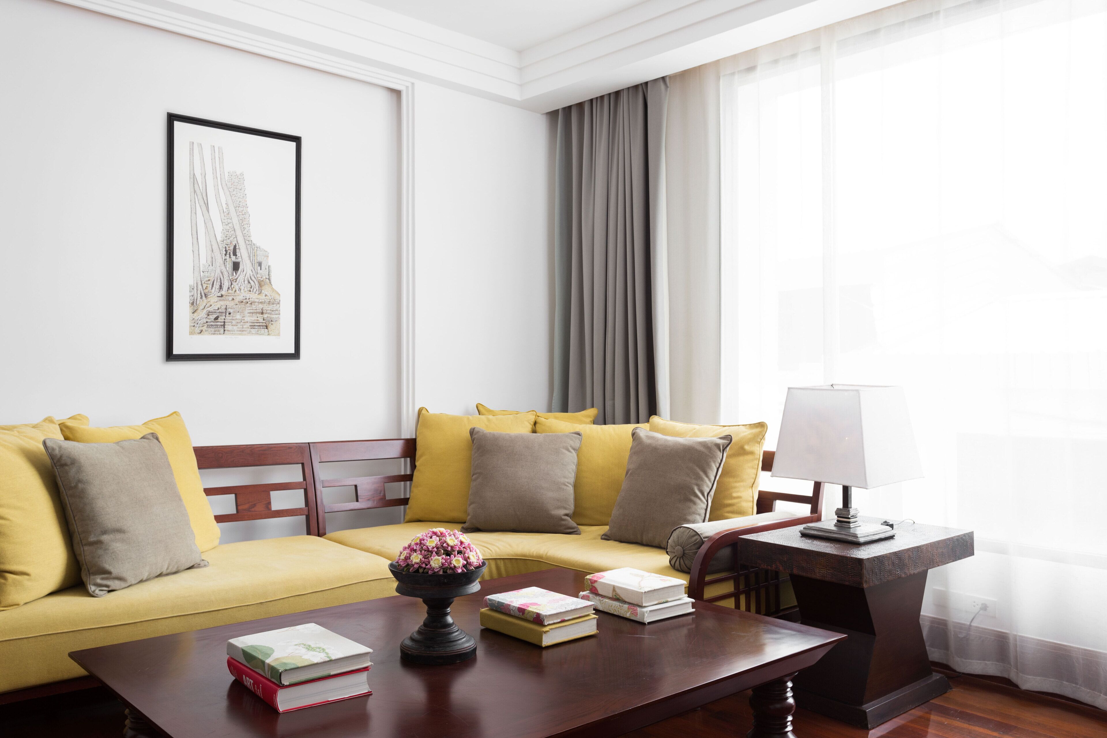 Una habitación de hotel moderna con un sofá amarillo, una mesa de centro de madera con libros y un florero, una lámpara y un cuadro en la pared.