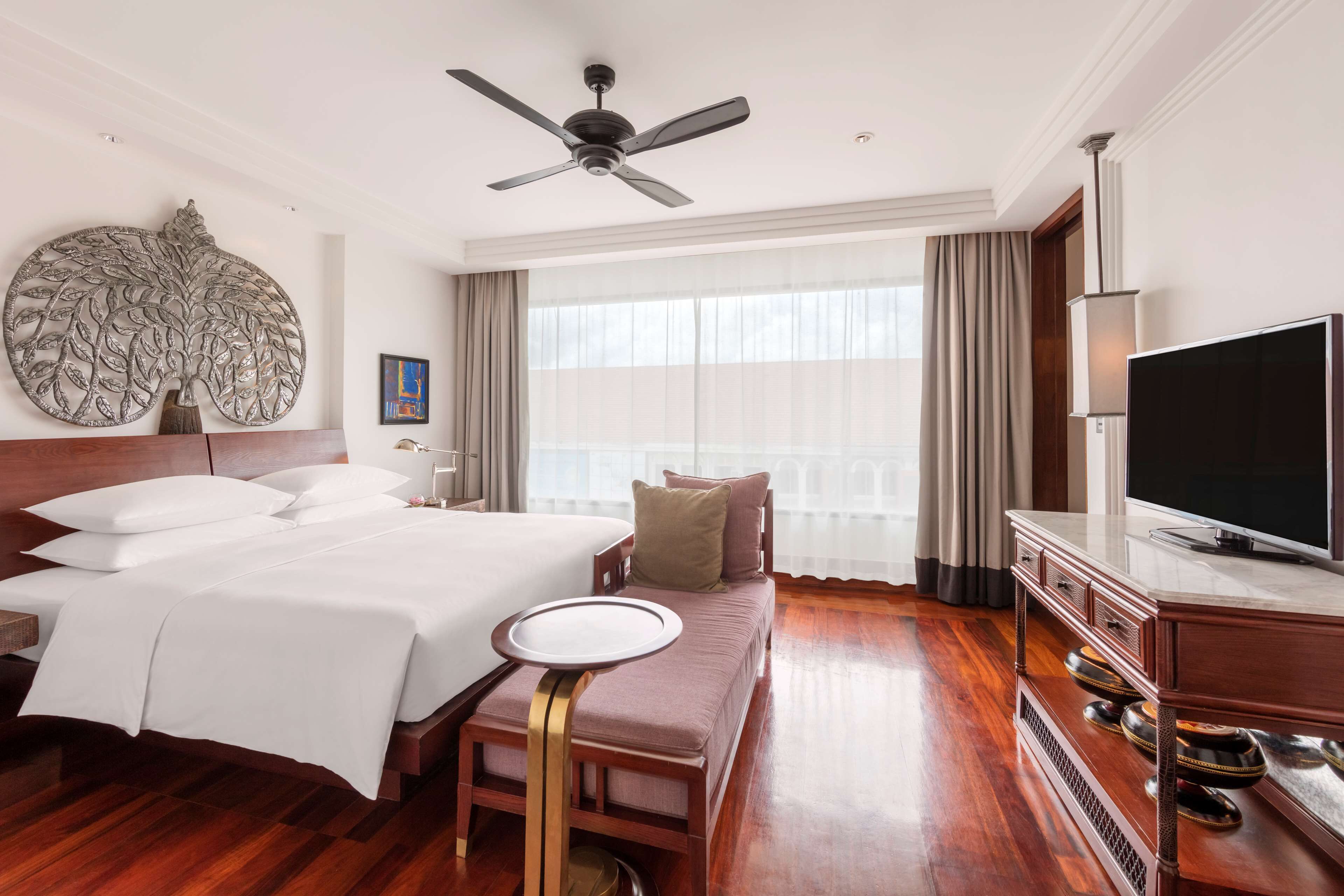 A hotel room with a large bed, a flat-screen TV, a ceiling fan, and a large window with curtains.