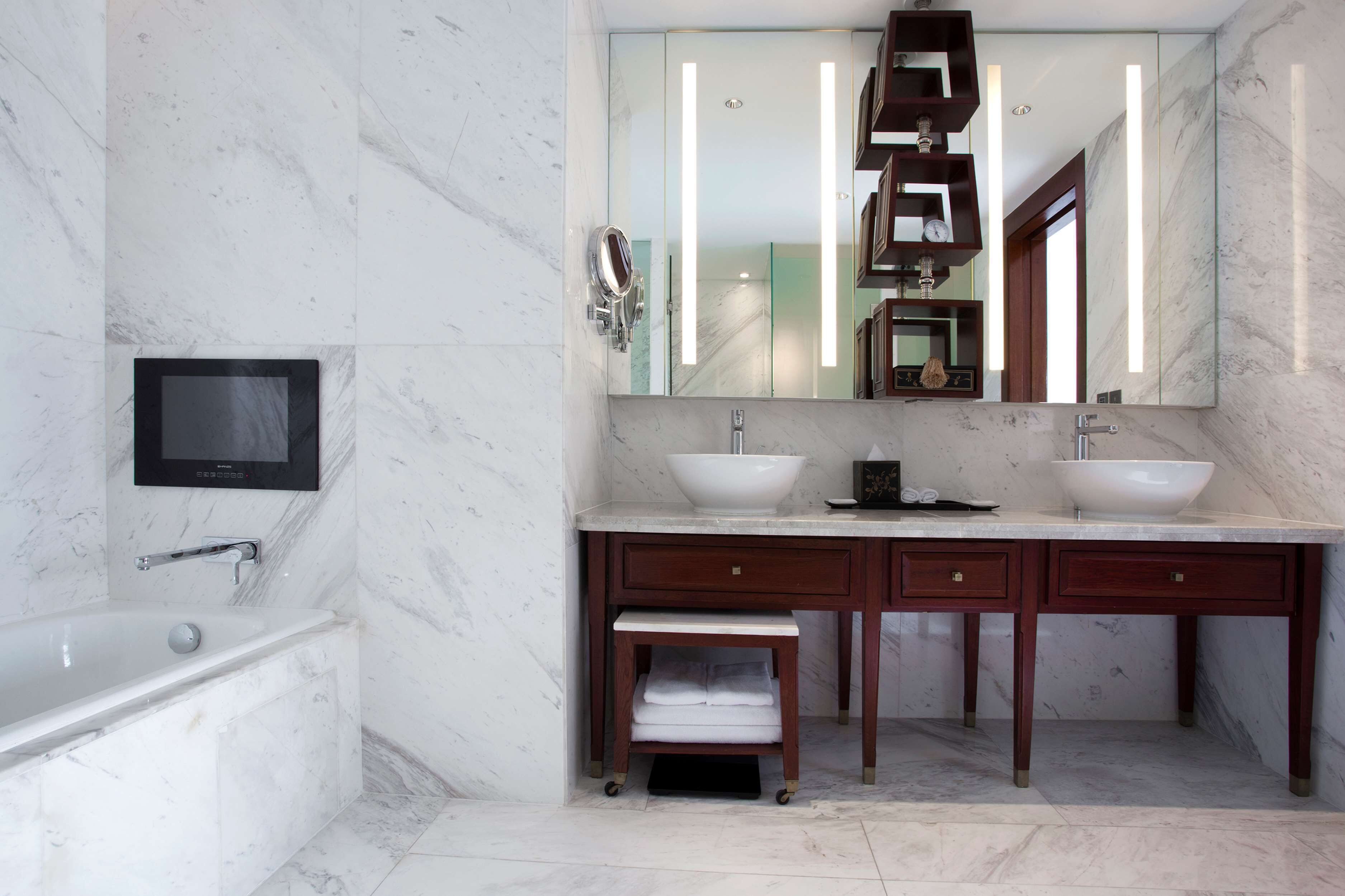 Un baño moderno con bañera, dos lavamanos, un mueble de tocador con cajones, un taburete pequeño y un televisor montado en la pared.