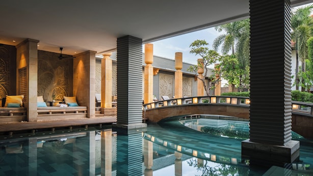 Un espace piscine intérieure paisible, agrémenté d’un pont en pierre, de coins salon au bord de la piscine et où se reflète la verdure environnante.
