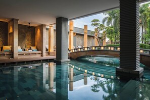 Ein ruhiger Innenpoolbereich mit Steinbrücke, Sitzgelegenheiten am Pool und Spiegelungen der umliegenden Bepflanzung.
