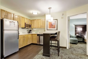 A compact kitchen with wooden cabinets, a refrigerator, microwave, and a dining area with a table and chairs. - Staybridge Suites O'Fallon Chesterfield by IHG (O'Fallon)