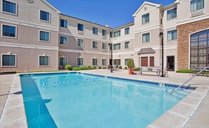 Una piscina con agua azul cristalina, rodeada por un patio con mobiliario exterior y un edificio con varias ventanas al fondo.