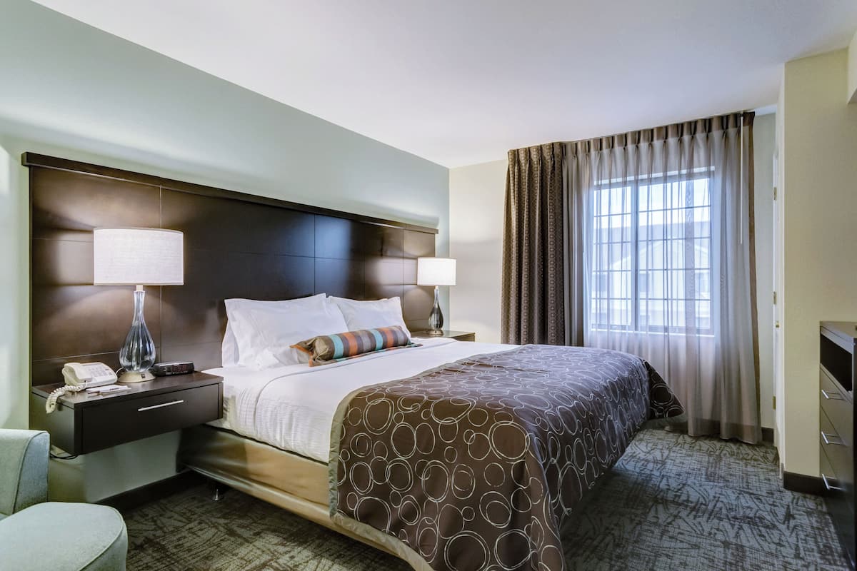 A hotel room with a large bed, two bedside tables with lamps, a nightstand, a chair, and a window with curtains.