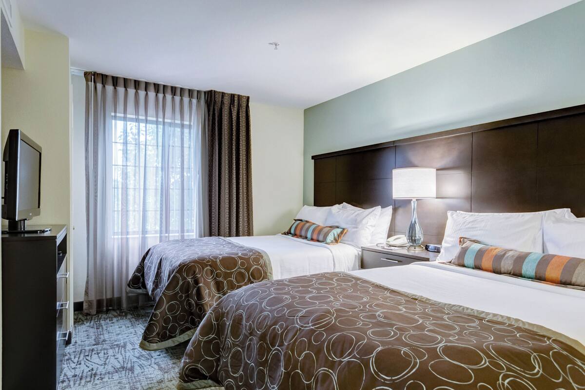 A hotel room with two beds, a desk with a lamp, a television, and a window with curtains.