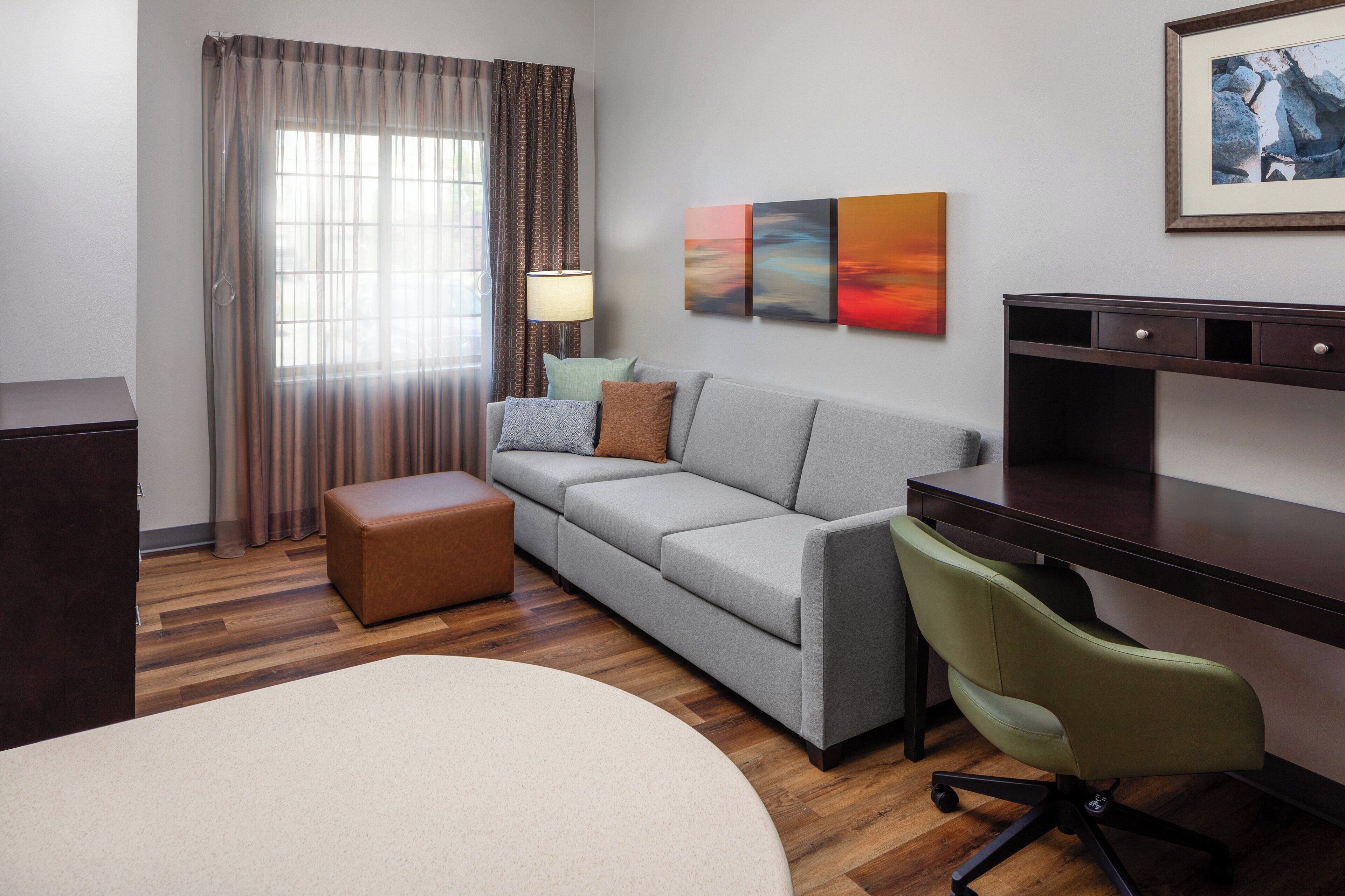 A hotel room with a sofa, a desk, a chair, a lamp, a painting, and a window with curtains.