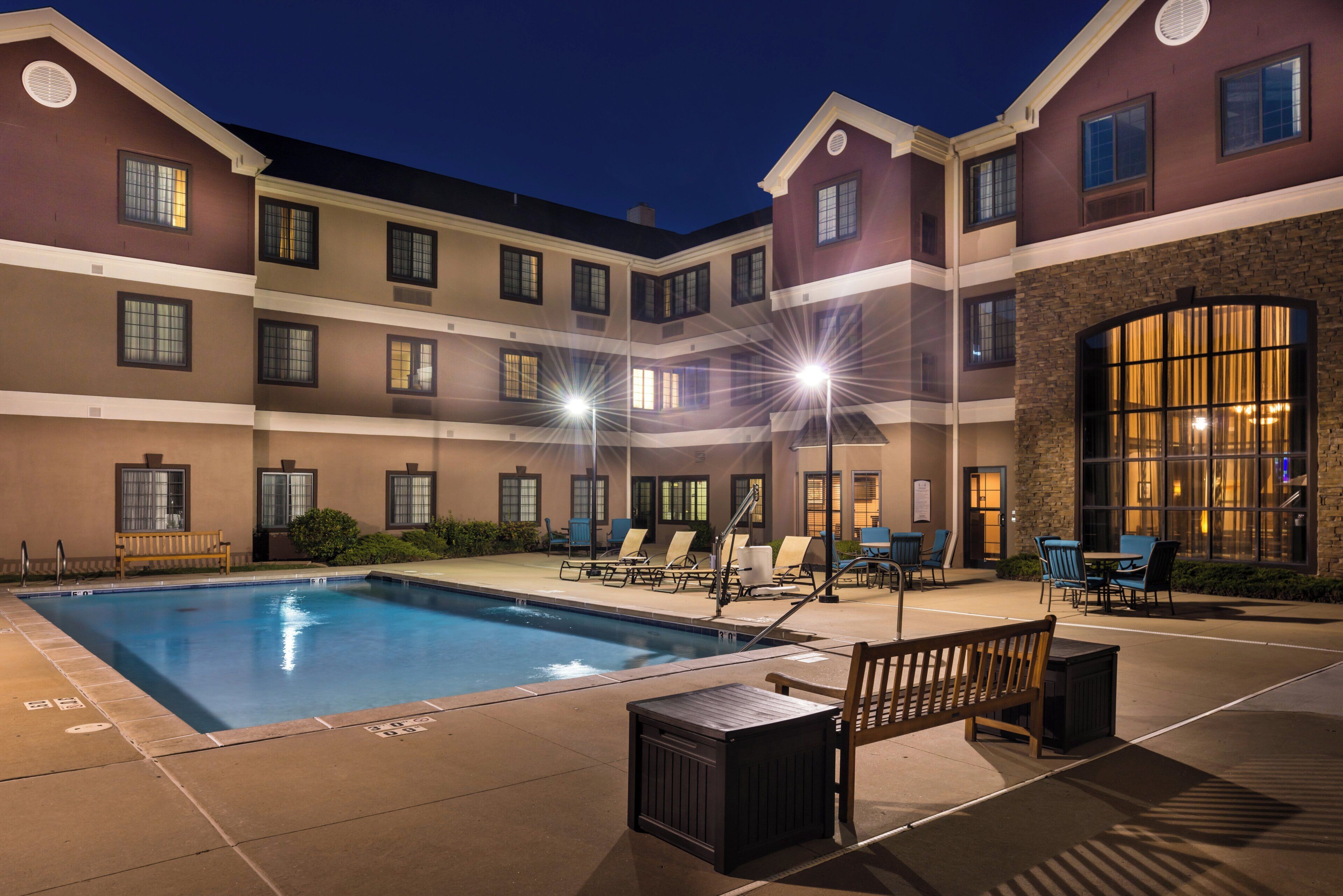 A hotel pool area with a rectangular pool, multiple lounge chairs, and a wooden bench.