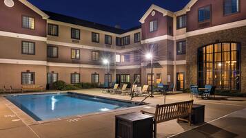 A hotel pool area with a rectangular pool, multiple lounge chairs, and a wooden bench.