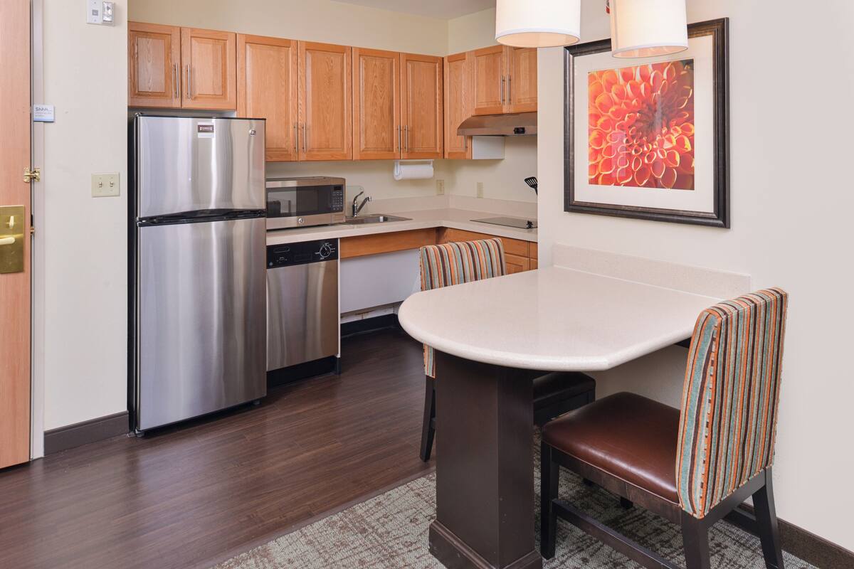 A modern kitchen with wooden cabinets, stainless steel appliances, a dining area with striped chairs, and a framed floral artwork on the wall.