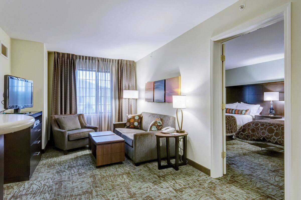 A hotel room with a sofa, armchair, bedside table, lamp, and a television.