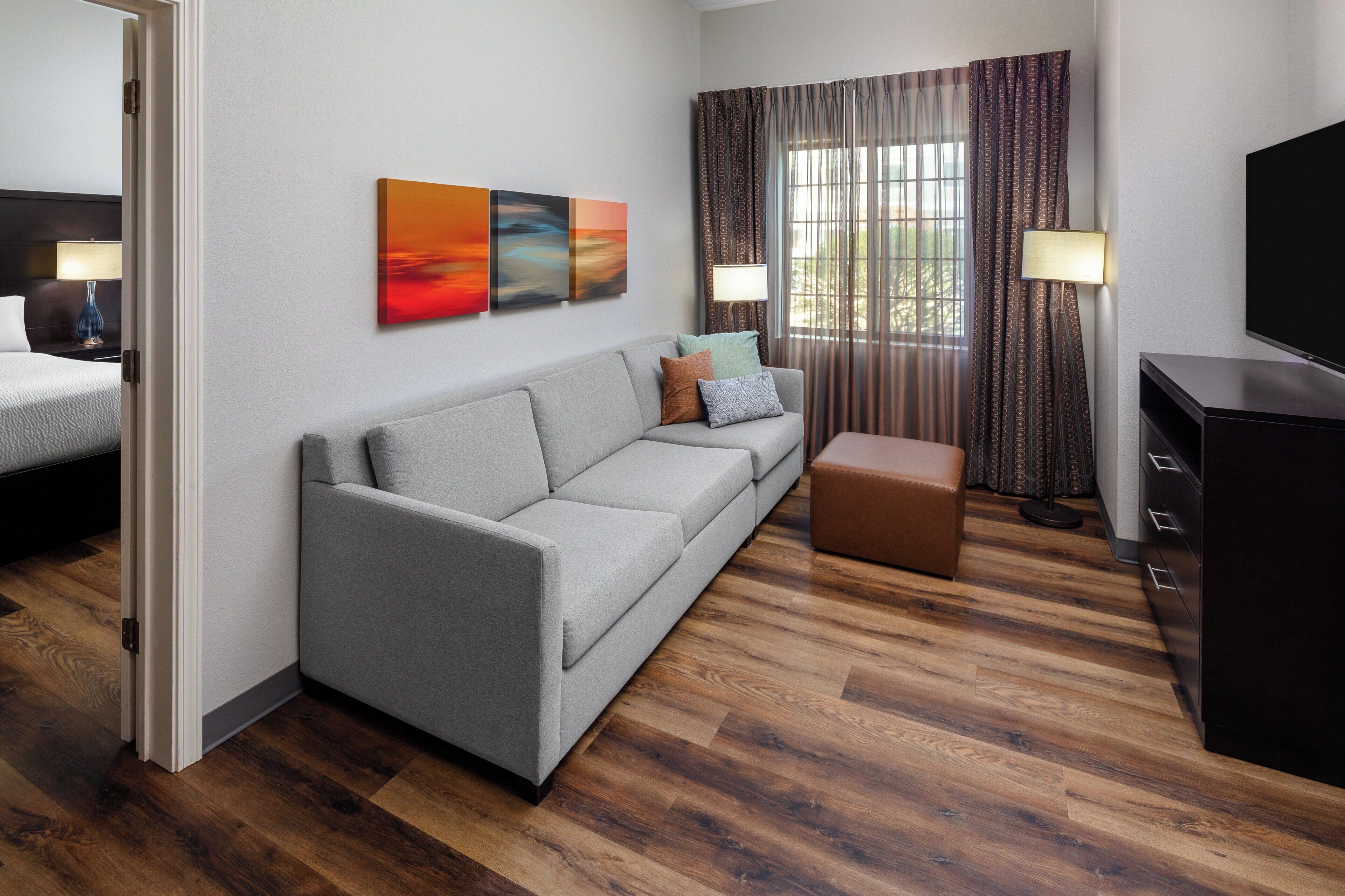 A modern hotel room with a grey sofa, a small ottoman, a TV stand, and a window with curtains.