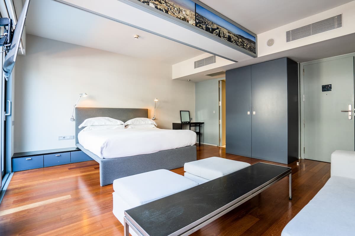 A modern hotel room with a bed, bedside table, and a desk with a chair. There is a wardrobe and a door visible.