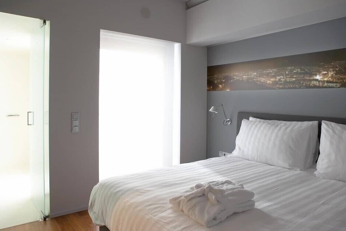 A modern hotel room with a large bed, a cityscape view, and a glass door leading to a balcony.