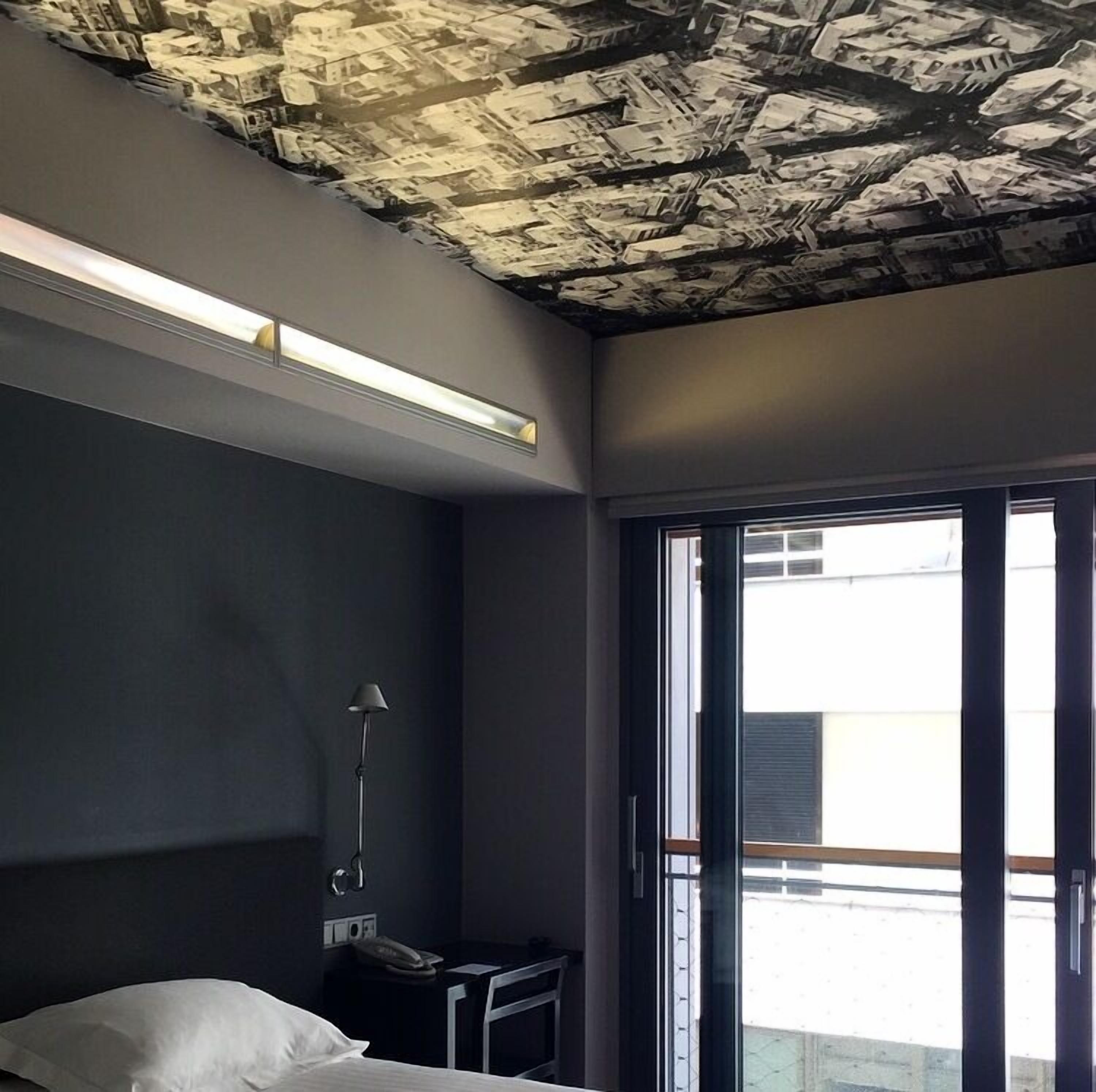 A modern hotel room with a dark color scheme, a bed, a desk with a lamp, and a ceiling with a unique textured design.