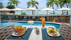 Un área de comedor junto a la piscina con vista al océano, palmeras y sillones de descanso.