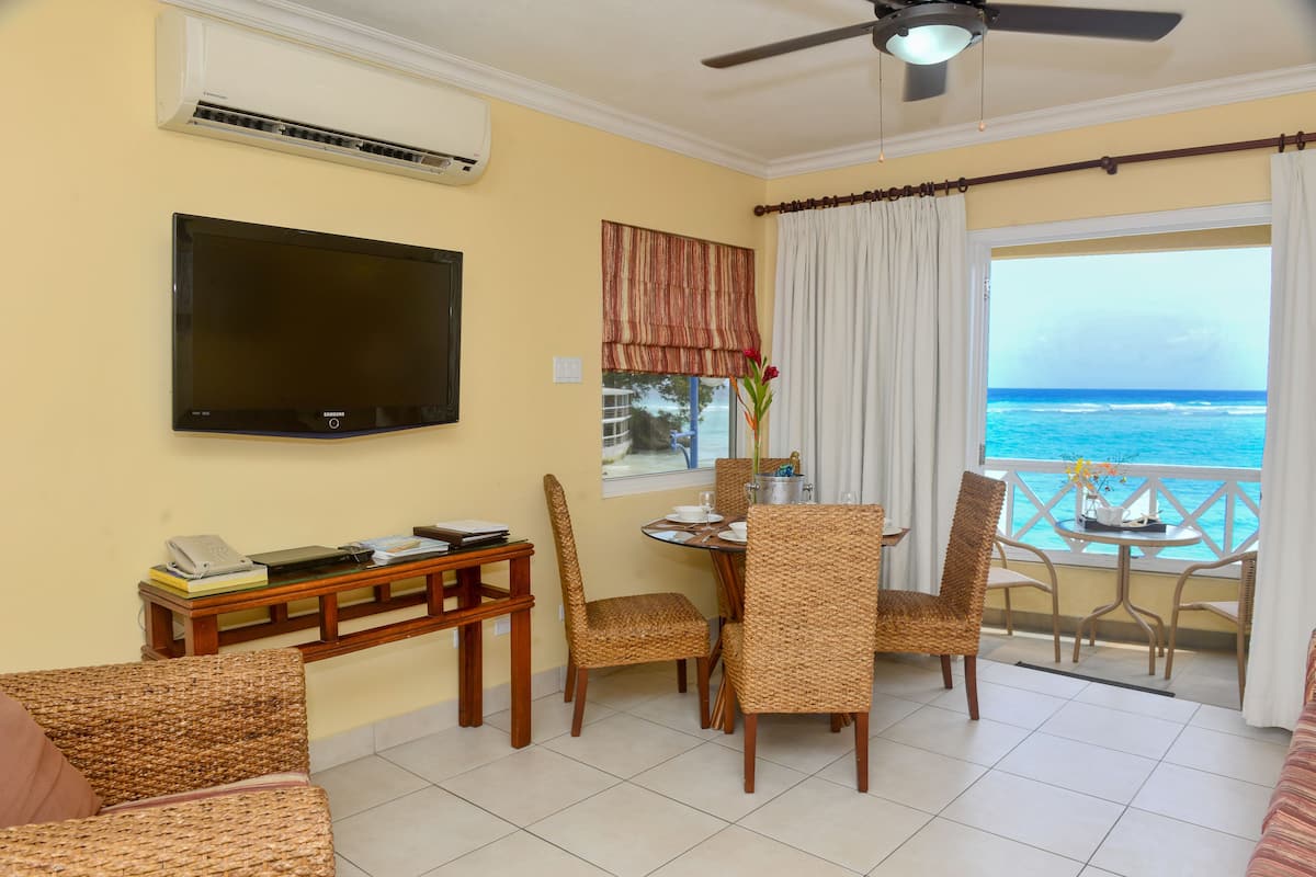 A living room with a TV, a dining table with chairs, a sofa, and a balcony with a view of the ocean.