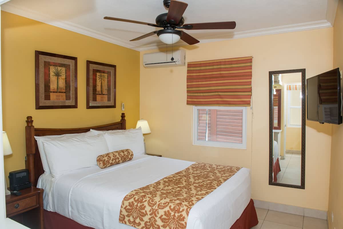 A hotel room with a bed, a ceiling fan, an air conditioning unit, a window with blinds, a mirror, and a television.
