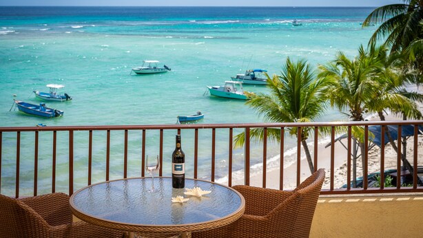 Un balcón con mobiliario de mimbre, una mesa con botella y vaso, y vista al mar con barcos y palmeras.