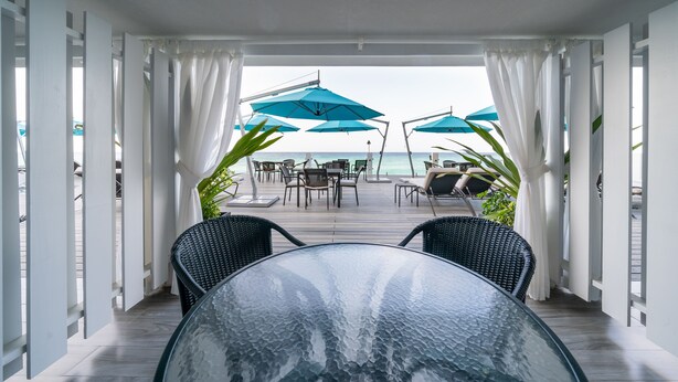 Mesa de vidrio con sillas en un espacio exterior cubierto con cortinas blancas y sombrillas azules, con vista a una playa.