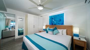 Une chambre d’hôtel comprenant un lit, un ventilateur de plafond, une table de chevet, une commode, une fenêtre avec des rideaux, un tableau décoratif au mur et un plancher en bois.