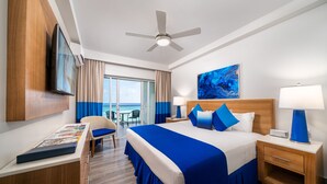 Habitación de hotel con cama, escritorio, silla, televisor, ventilador de techo y ventana con vista al océano.