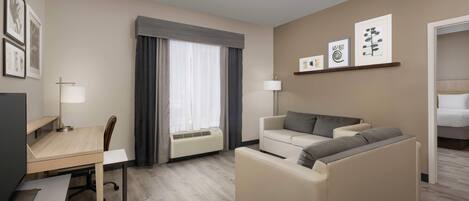 Una habitación de hotel con escritorio, silla, sofá y cama. Hay cuadros en la pared, una ventana con cortinas y un equipo de aire acondicionado.