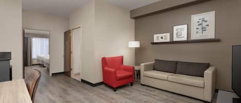 A hotel room with a wooden floor, a red armchair, a beige sofa, a TV, and framed artwork on the wall.