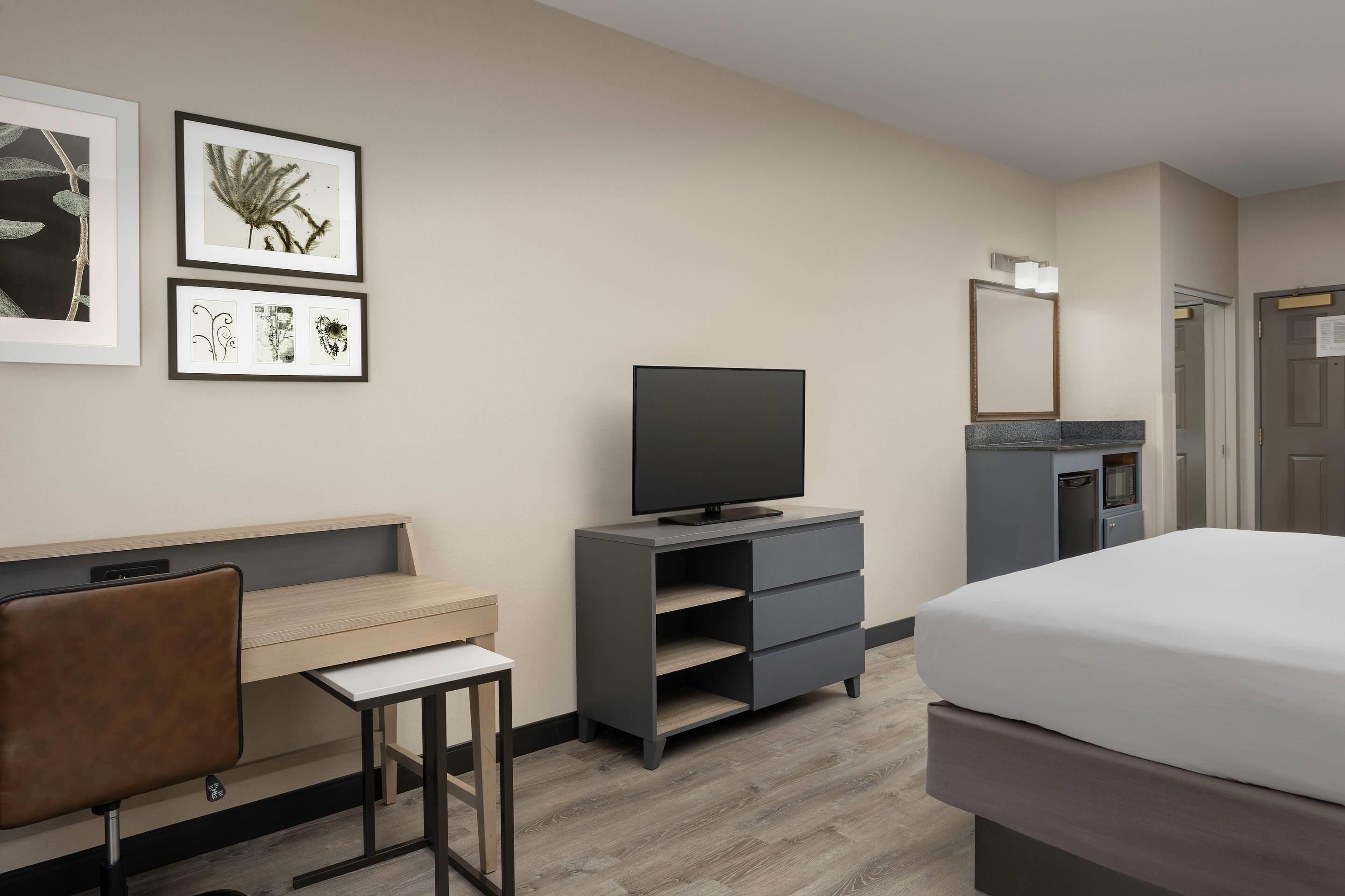 Une chambre d’hôtel comprenant un lit, un bureau avec une chaise, une télévision, une commode et des œuvres d’art botanique encadrées accrochées au mur.