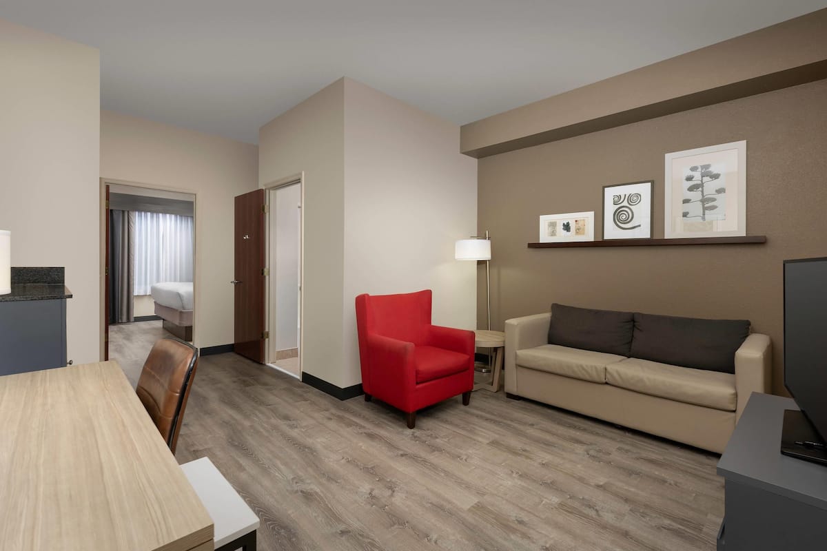A hotel room with a wooden floor, a red armchair, a beige sofa, a TV, a lamp, and framed artwork on the wall.