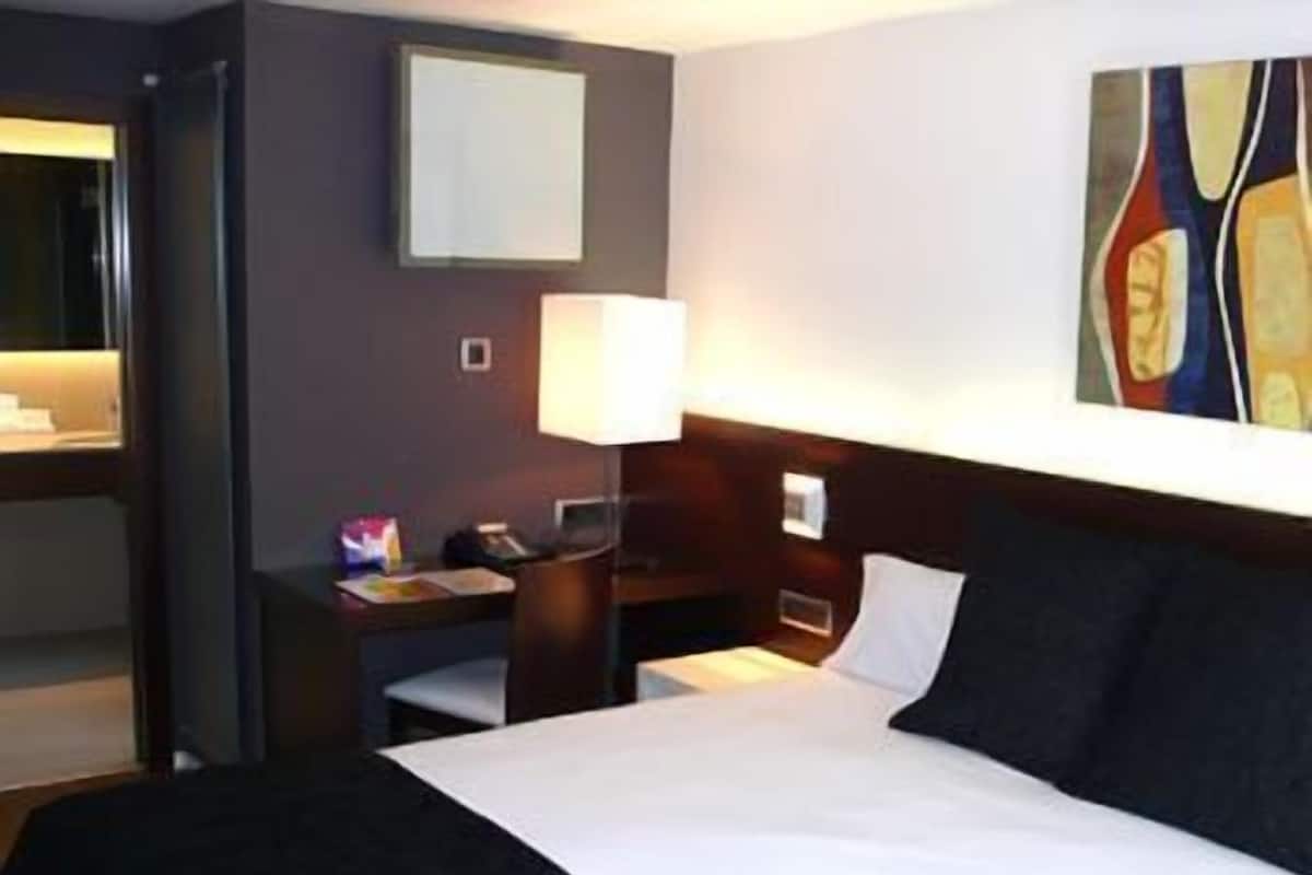 A hotel room with a bed, a desk with a lamp, a chair, a small table, and a painting on the wall.