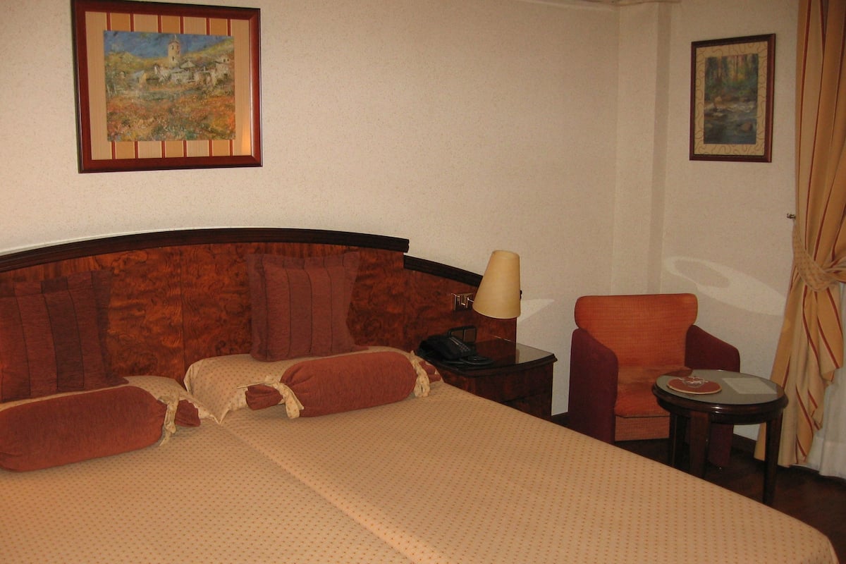 A hotel room with a large bed, a wooden headboard, a desk with a lamp, a chair, and two paintings on the wall.