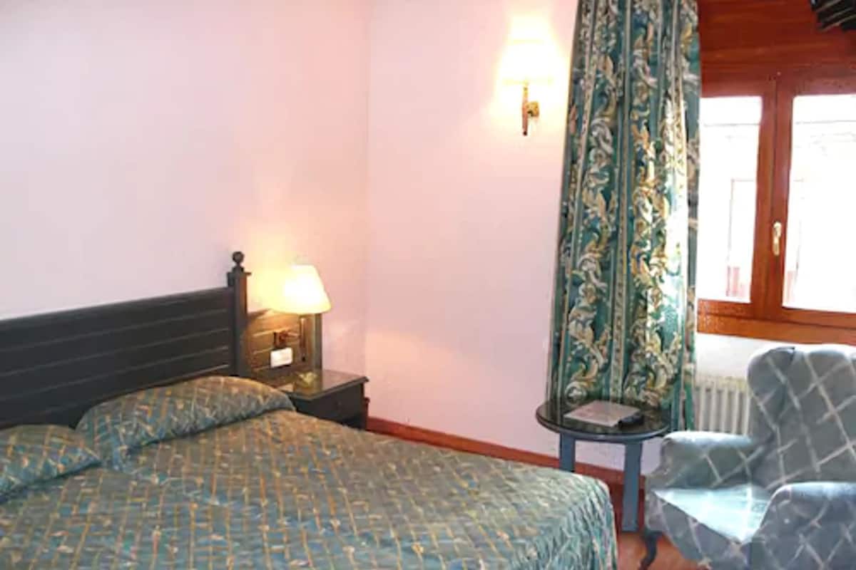 A bedroom with a bed, a nightstand with a lamp, a chair, and a window with curtains.