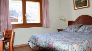 Une chambre à coucher comprenant un lit, une chaise, un bureau, une fenêtre avec des rideaux, une table de chevet, une lampe et un tableau encadré accroché au mur.