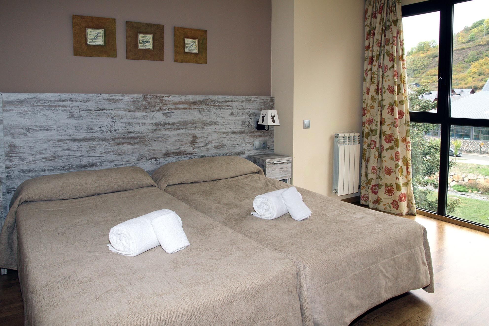 A double bed with two rolled towels, a nightstand with a lamp, and a view of greenery through a window.
