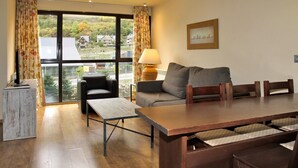 A cozy living room with a sofa, a wooden table, and a chair. There is a window with curtains, a lamp, and a framed picture on the wall. - Aparthotel La Vall Blanca (Vielha e Mijaran)