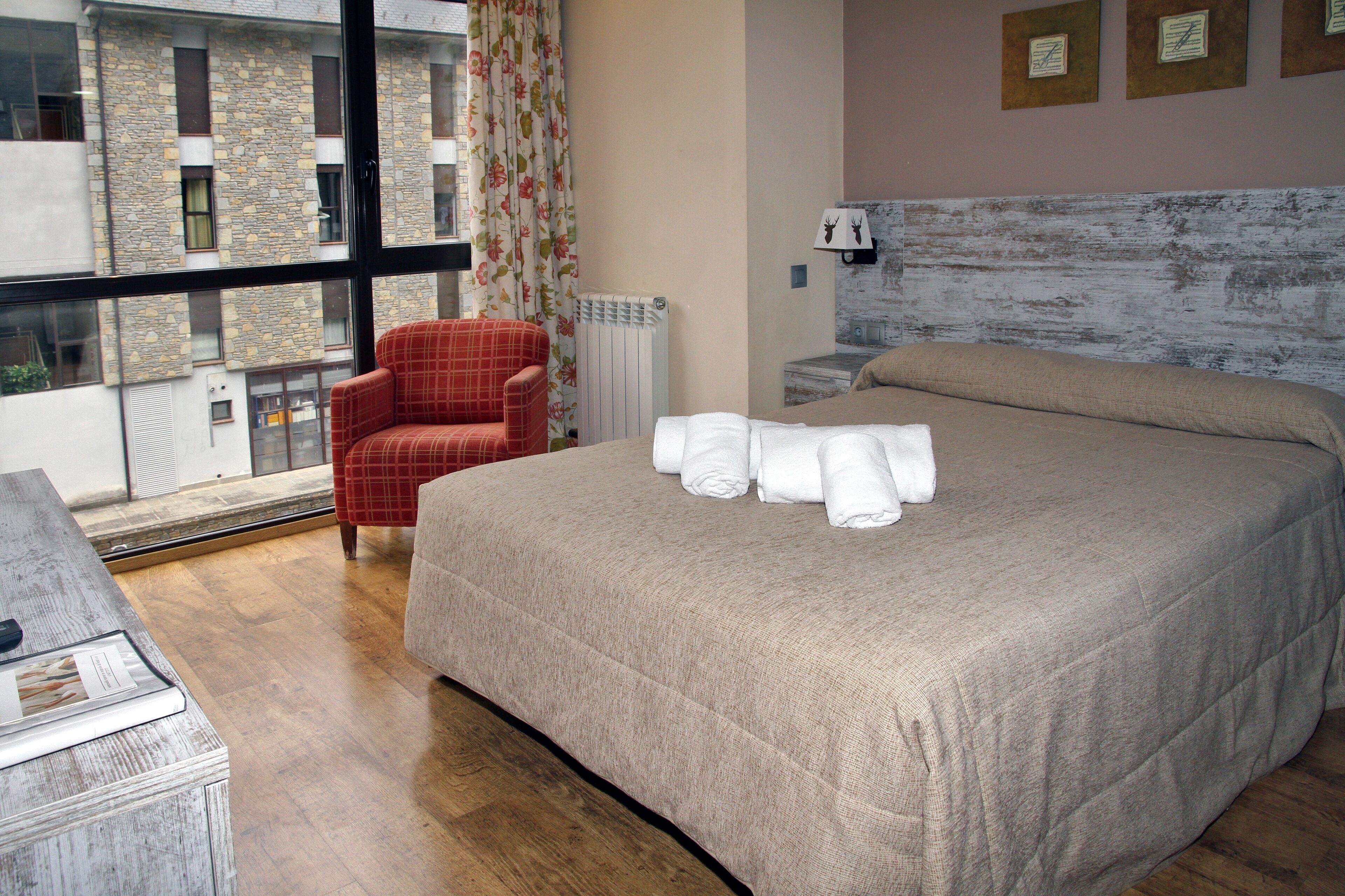 A bedroom with a large bed, a red plaid armchair, a radiator, and a window with a view of a building.
