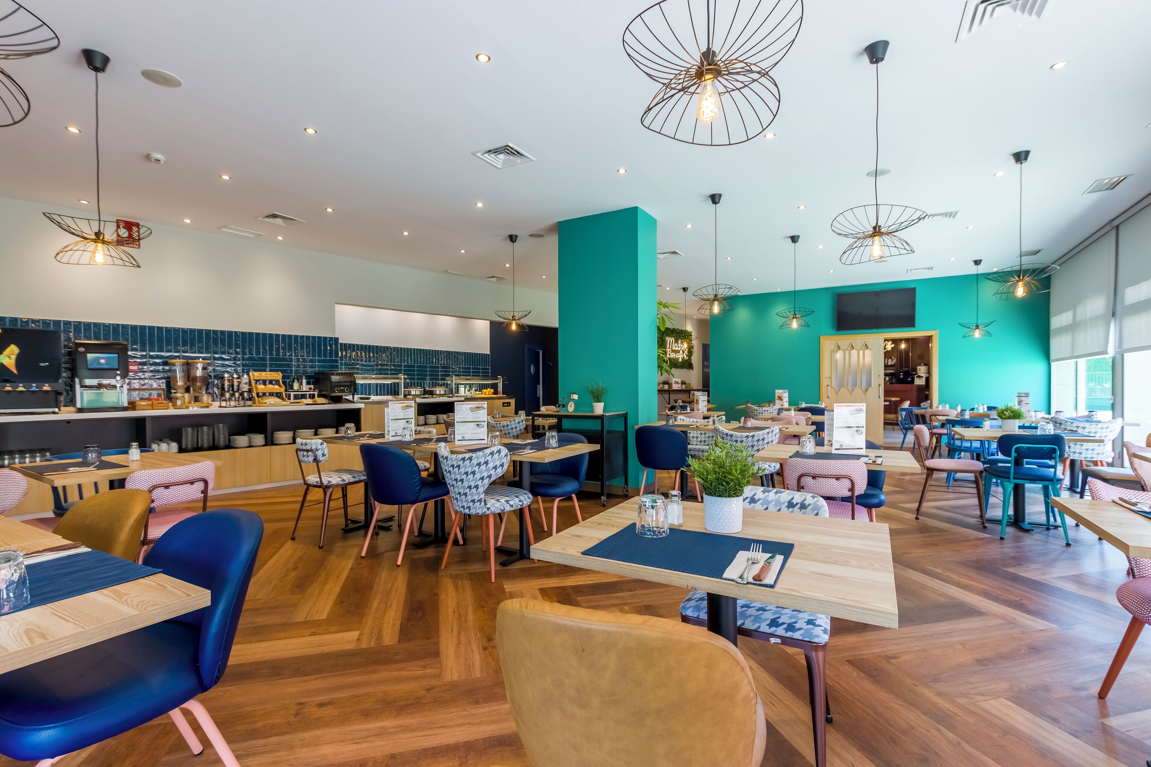Een modern restaurant met houten vloeren, wanden in blauwgroen en verschillende zitmogelijkheden, zoals tafels en stoelen.