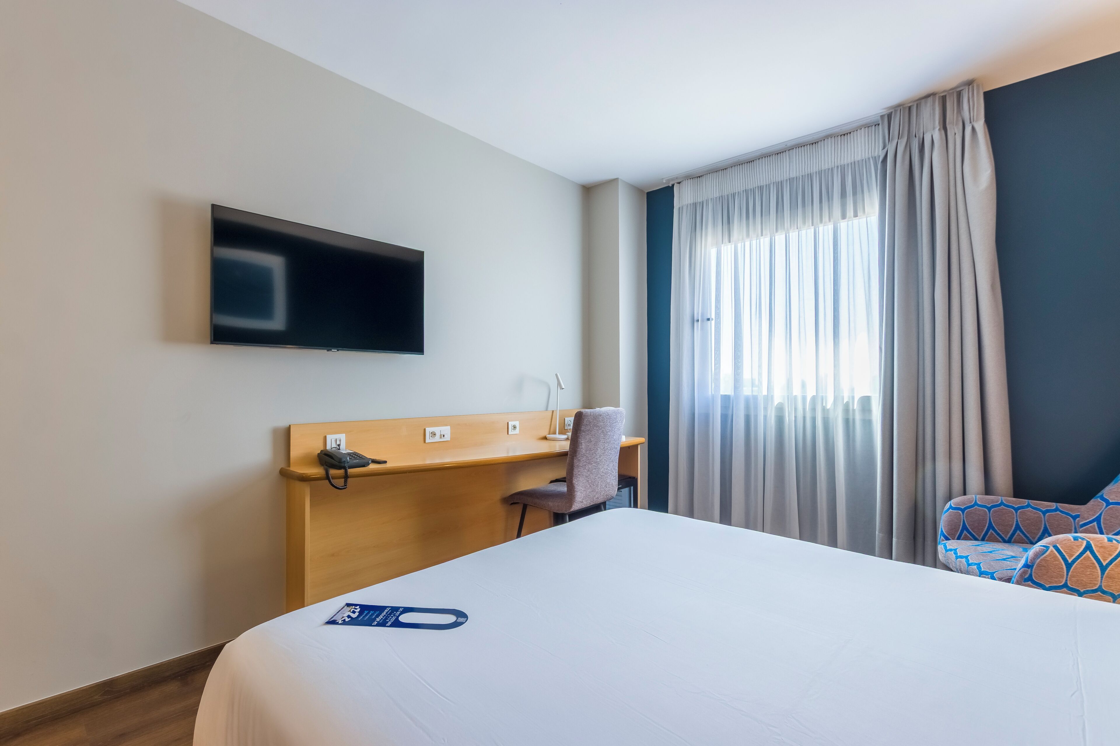 Una habitación de hotel con cama, escritorio con teléfono, silla, televisor montado en la pared y ventana con cortinas.
