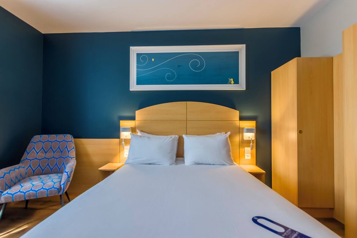 A hotel room with a large bed, a chair, a wooden wardrobe, and a wall-mounted artwork.