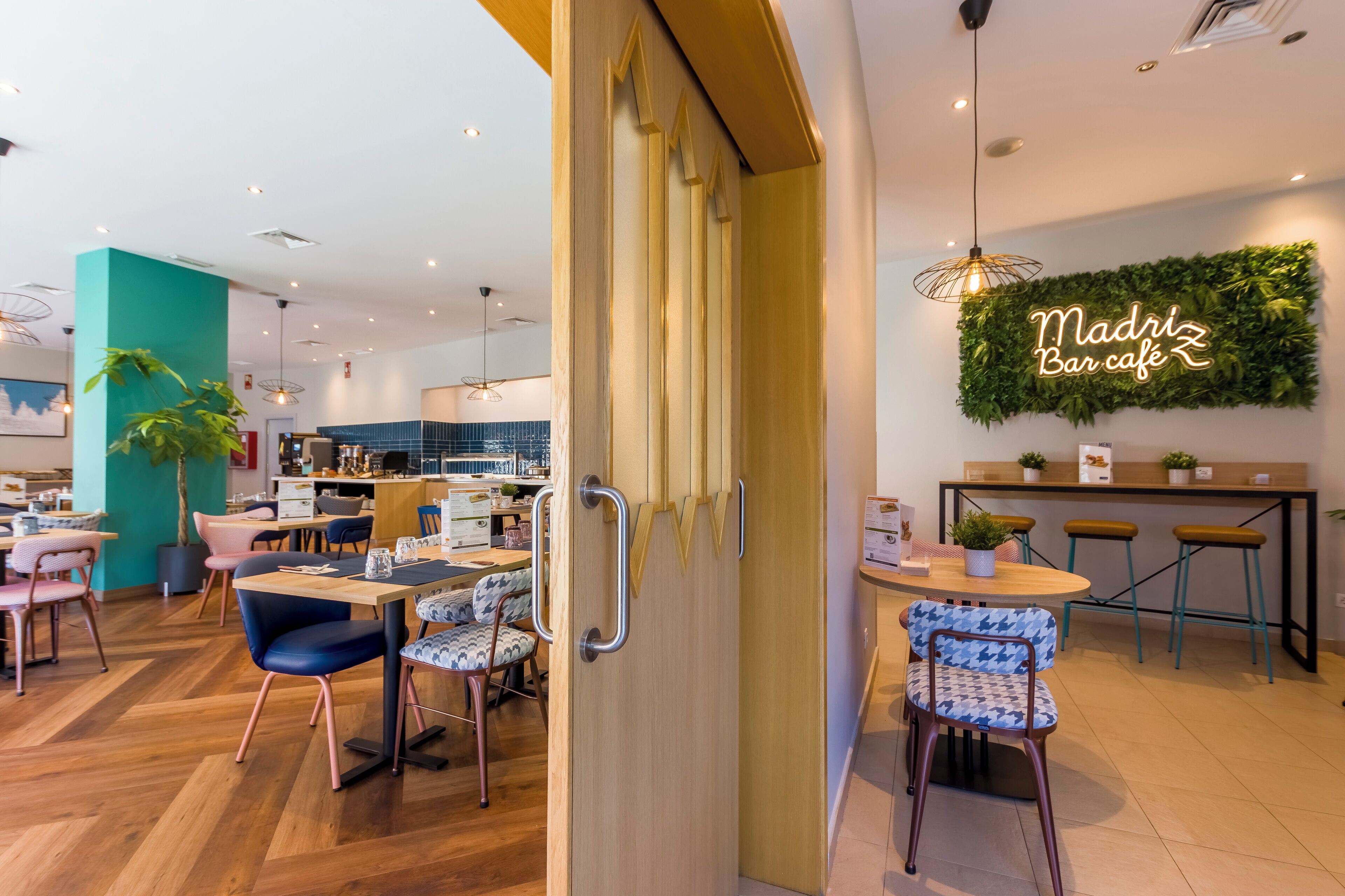 Un intérieur moderne avec un plancher en bois, un coin bar avec des tabourets et un panneau néon indiquant « Madriz Bar café ».