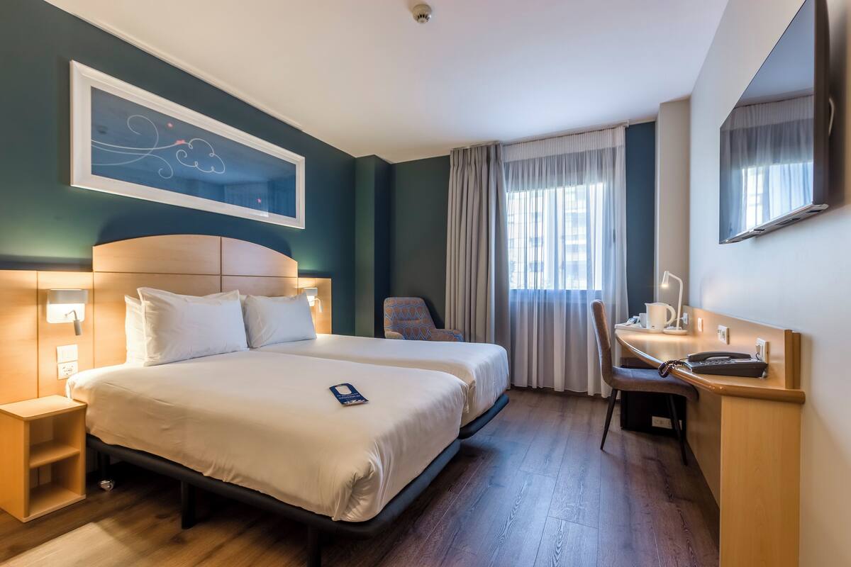 A hotel room with two beds, a desk, a chair, a nightstand, and a large window with curtains.