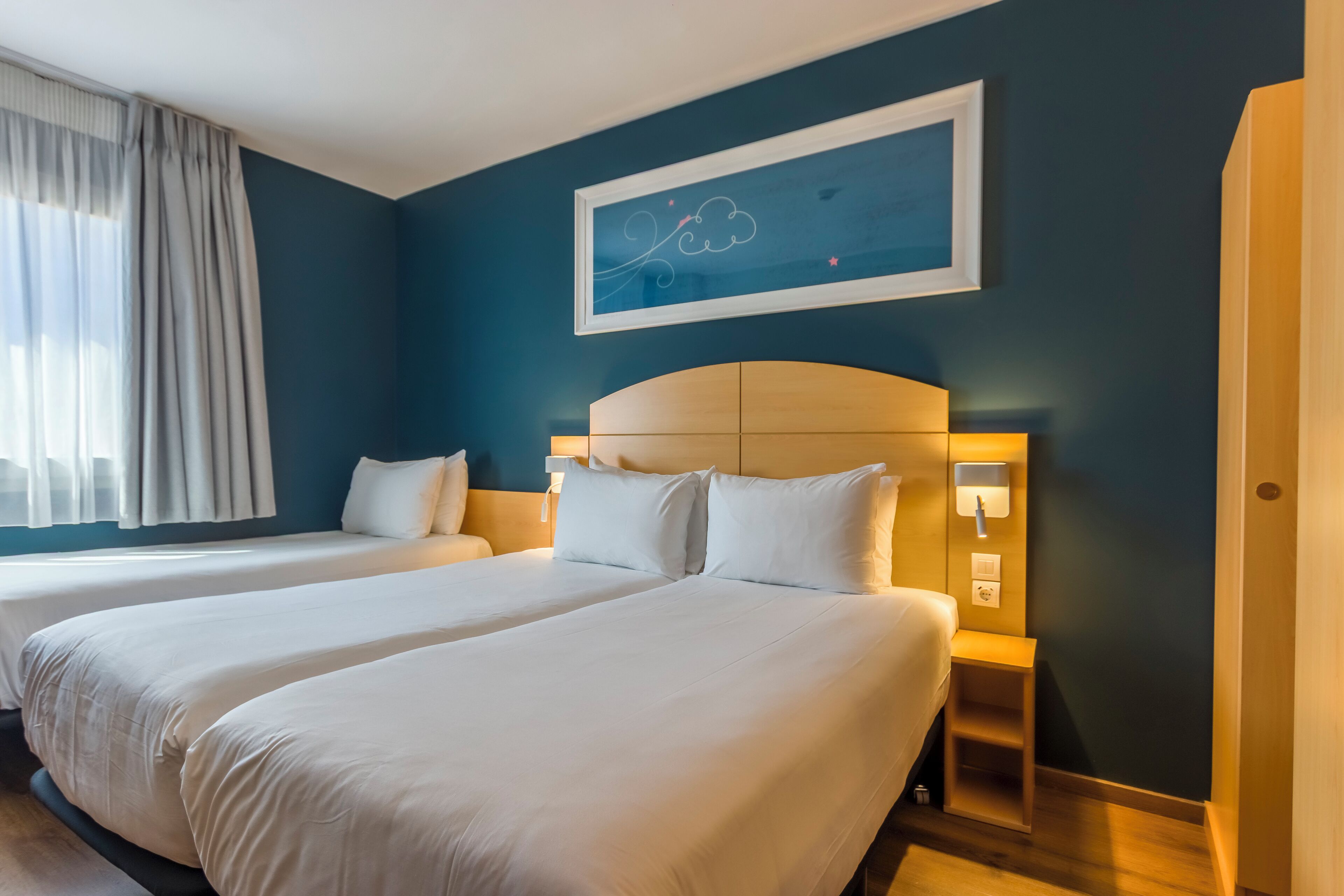 Quarto de hotel com duas camas, uma janela grande com cortinas, uma obra de arte na parede e uma mesa de cabeceira com abajur.